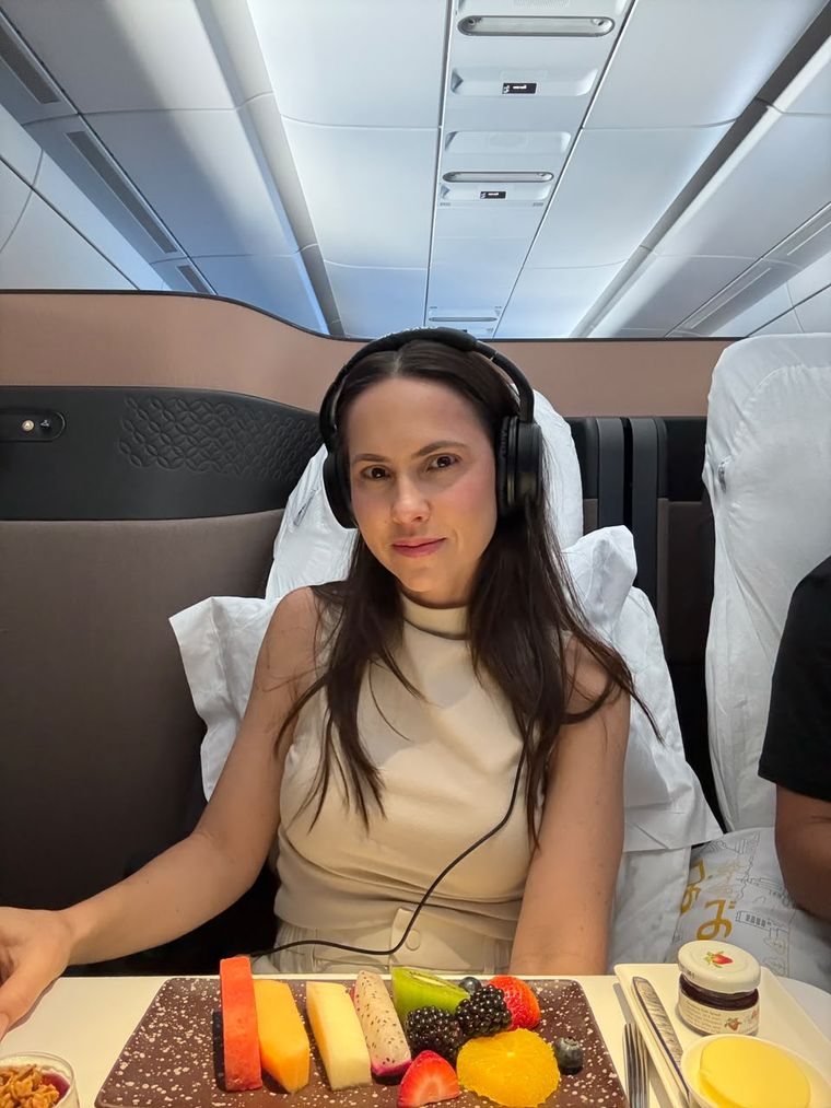 Miriã sentada na poltrona de um avião, com fones e um prato de frutas